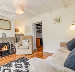 A living room with a beige armchair and sofa with cushions a lit fireplace a lamp and a patterned rug at Vi's Valley View in Pentewan