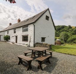 An outdoor seating area with wooden picnic benches and a white stone house with windows and chimneys at Penarth in Cregrina near Builth Wells