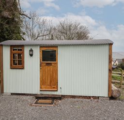 A small corrugated metal hut with a wooden door and window on a gravel surface at Rabbit Hill Hut in Govilon