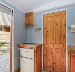 A small kitchen area with a fridge, wooden cabinets, and a door at Rabbit Hill Hut in Govilon