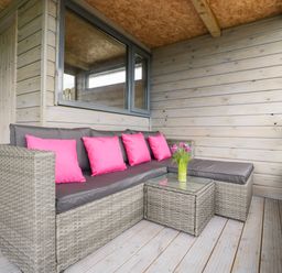 An outdoor seating area with a wicker sofa with gray cushions and pink pillows and a small glass table with flowers at Thornhills Lodge Othery near Middlezoy