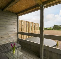 A covered wooden patio with a wicker sofa and glass table with flowers overlooking a field at Thornhills Lodge in Othery near Middlezoy