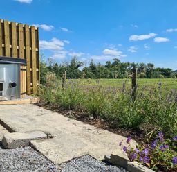 A garden with a wooden hot tub on a paved area next to a flower bed and a green field at Thornhills Lodge in Othery near Middlezoy