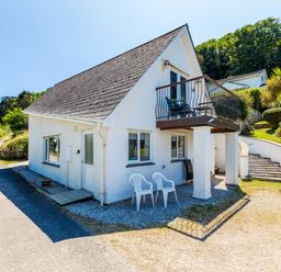 A white house with a balcony and plastic chairs outside near a curved staircase and trees at Baywatch Sands Polzeath