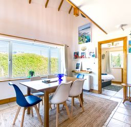 A dining area with a wooden table and six chairs next to a large window with hedges outside and view into a bedroom at Baywatch Sands in Polzeath