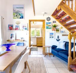 A dining area with a wooden table and chairs next to a blue sofa and wooden staircase at Baywatch Sands in Polzeath
