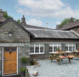 An outdoor patio area with wooden picnic tables and a stone building with a wooden door and multiple windows at Thorney How in Grasmere