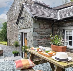 An outdoor wooden picnic table with a hat cushion plants and food outside a stone house at Thorney How in Grasmere
