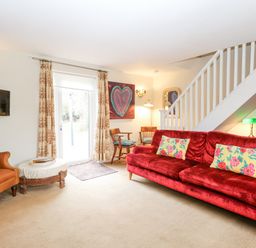 A living room with a red sofa and floral cushions an orange armchair and wooden chairs near a staircase at Willow Cottage in Hickling near Catfield