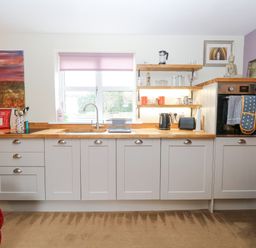 A kitchen with white cabinets wooden countertops a window with a pink blind and shelves with kitchen items at Willow Cottage in Hickling near Catfield