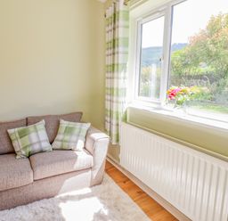 A living room with a beige sofa with green and white plaid cushions by a window with green plaid curtains and a vase of flowers on the windowsill at Cam O'r Fechan Annexe in Llangollen