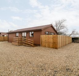 A single story lodge with wooden exterior and a ramp surrounded by gravel and wooden fencing at Bluebell Lodge Meadow view lodges in Brean