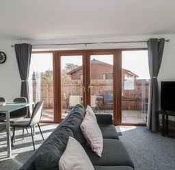 A living room with a gray sofa and cushions a glass dining table with chairs a wall clock and a tv on a stand with glass doors leading to a fenced patio at Bluebell Lodge Meadow view lodges in Brean