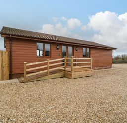 An exterior view of a single story wooden building with a sloped roof and a wooden ramp surrounded by gravel at Delphine Lodge Meadow View Lodges in Brean