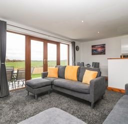 A living room with gray sofas and orange cushions near glass doors overlooking a green field at Delphine Lodge Meadow View Lodges Brean