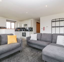 A living room with gray sofas and yellow and white cushions next to a kitchen with white cabinets and black backsplash at Delphine Lodge Meadow View Lodges in Brean