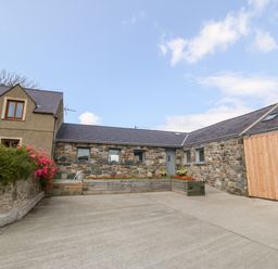 An exterior view of a stone and wooden house with flower beds and a concrete driveway at Y Beudy Dinas near Bontnewydd