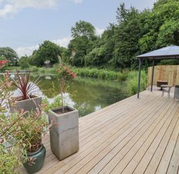 A wooden deck with potted plants a gazebo chairs and a small table next to a pond with trees at Maple Lodge at Bridge Lake Farm & Fishery in Chacombe