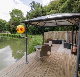 A patio area with chairs and a table near a pond at Maple Lodge at Bridge Lake Farm & Fishery Chacombe