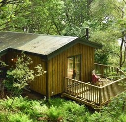 A wooden cabin with a deck and chairs surrounded by trees and greenery at Chapel House in Torridon