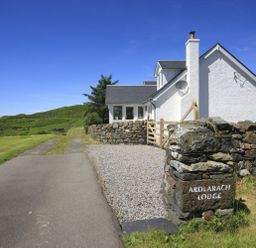 A white house with a chimney next to a stone wall and a gravel driveway at Ardlarach Lodge Cullipool Isle Of Luing