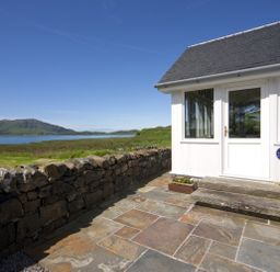 Stone patio with steps leading to white door and windows next to stone wall with grassy landscape and water view at Ardlarach Lodge in Cullipool Isle Of Luing