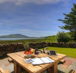 A wooden picnic table with fruit, wine, and maps on a patio overlooking a grassy area and stone wall with water and hills in the background at Ardlarach Lodge in Cullipool Isle Of Luing