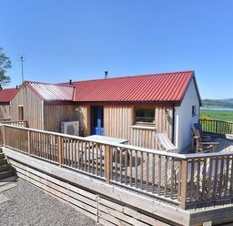 A wooden house with a red roof and blue door with a fenced deck overlooking fields and water at Skylarks Ferintosh near to Dingwall
