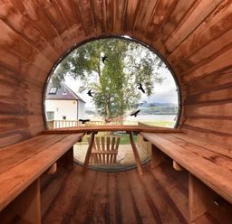 An interior view of a wooden barrel-shaped room with benches on both sides and a round window looking out to a tree and a house at Skylarks in Ferintosh near to Dingwall