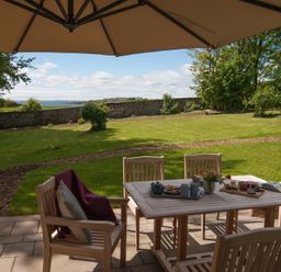 A patio with wooden table and chairs under an umbrella overlooking a garden with a stone wall and trees at Beltie Lodge in Borgue