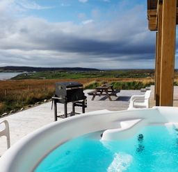 An outdoor wooden deck with a hot tub white chairs a barbecue grill and a picnic table overlooking a grassy landscape and water at Eagle Lodge in Uig near Stornoway