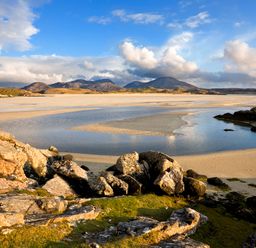 Rocky foreground with patches of grass leading to a sandy beach and a body of water with mountains in the background at Eagle Lodge Uig near Stornoway