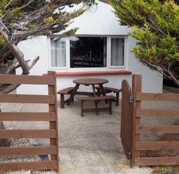 An outdoor area with a round wooden picnic table and benches behind a wooden gate and fence in front of a white building with a window Braemore Chalet Tarbert Isle Of Harris