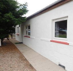 A white exterior wall of a building with windows and a door next to a gravel and concrete path with trees on the side at Braemore Chalet Tarbert Isle Of Harris