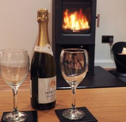 A bottle of sparkling wine and two empty glasses on a wooden table with a lit fireplace in the background at Braemore Chalet in Tarbert Isle Of Harris