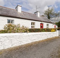 A white stone cottage with red doors surrounded by a white stone wall and bushes at Cartron Cottage in Ballintubber County Mayo