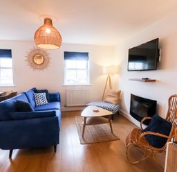 A living room with a blue sofa a patterned chair a rattan chair a coffee table a wall-mounted tv and two windows at Stoker's Rest in Whitby