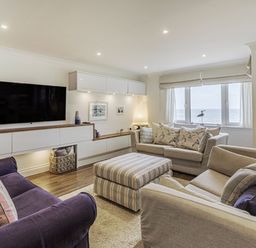 A living room with sofas a striped ottoman a wall-mounted television and a window overlooking the sea at Apartment 18 Burgh Island Causeway in Bigbury-On-Sea