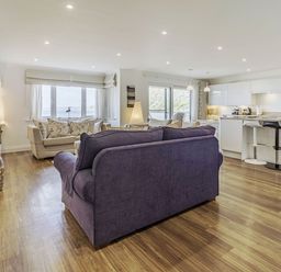 An open living room with a purple sofa and beige chairs facing a wall-mounted TV with a floating media console and a kitchen with white cabinets and bar stools at Apartment 18 Burgh Island Causeway in Bigbury-On-Sea