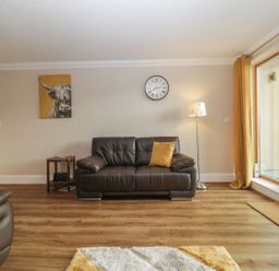 A living room with a brown leather sofa a side table with a radio a standing lamp a wall clock a yellow cushion a cow painting and large windows with yellow curtains at Townbridge Apartment in Weymouth