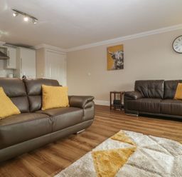A living room with brown leather sofas and yellow cushions a wooden floor a clock on the wall and a kitchen area at Townbridge Apartment in Weymouth