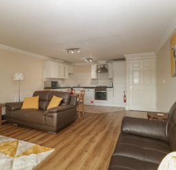 An open living room and kitchen area with brown sofas yellow cushions a wooden dining table and white kitchen cabinets at Townbridge Apartment in Weymouth