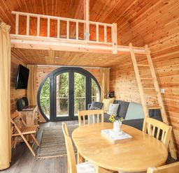 An interior of a wooden cabin with a dining table, chairs, a sofa, a ladder leading to a loft, and large circular doors at The Hideaway Pod in Rhoslan near Criccieth