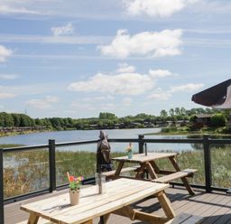 A wooden deck with picnic tables and flowers overlooking a lake with houses and trees at Parkers Place in Carnforth