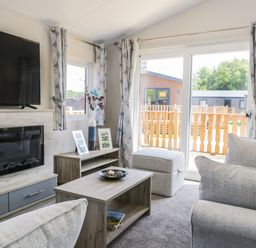 A living room with a television above a fireplace a wooden coffee table a small cabinet with vases and framed photos and sliding glass doors at Parkers Place in Carnforth