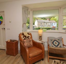 A sitting area with two chairs a small table with a lamp a basket with firewood and a window showing a house outside at Barleside in Dulverton