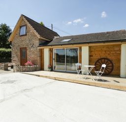 A cottage with a wooden deck table and chairs and a large wagon wheel on the wall at Steel Works Farm in Gorsley