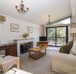 A living room with a white sofa green cushions a wooden coffee table a wooden desk and chair a fireplace and sliding glass doors at Manor Farm Lodge in Bowerchalke