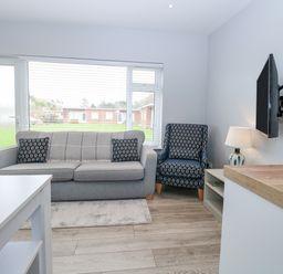 A living room with a grey sofa and patterned armchair a wall mounted TV and a lamp on a side table at 12 Siesta Mar in Mundesley