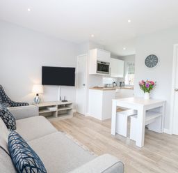A living area with a gray sofa, an armchair, a wall-mounted TV, a small dining table with stools, a lamp, and an open kitchen at 12 Siesta Mar in Mundesley